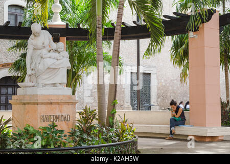 Una giovane donna controllando il suo telefono cellulare al cortile vicino alla statua descrive la maternità, Merida, Yucatan, Messico. Foto Stock