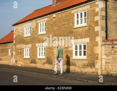 Vecchio / senior man walking: montare e rivestimento uomo anziano tenuto vivace, passeggiata quotidiana nella calda, mattino sunshine, passato una vecchia casa di pietra. Inghilterra, Regno Unito. Foto Stock