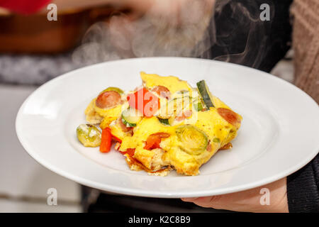 Porzione di frittata gustosa con salsiccia tagliata a fettine, verdure sulla piastra bianca Foto Stock