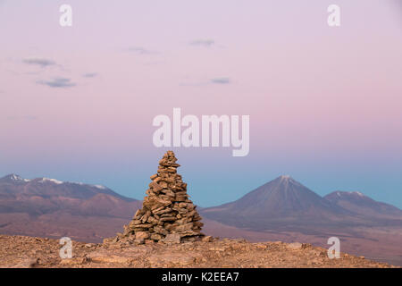 Apacheta / Apachita - una pietra cairn nelle Ande, un piccolo mucchio di pietre costruite lungo il sentiero in alta montagna con il vulcano Licancabur (5916 m sopra il livello del mare) e il vulcano Juriques all' estrema destra (5704 m) sullo sfondo al tramonto, il Deserto di Atacama, San Pedro, Cile Foto Stock