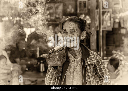 Felice e vecchia donna rugosa fuma una grande cheroot sigaro in pubblico. Vintage in bianco e nero elaborata. Foto Stock