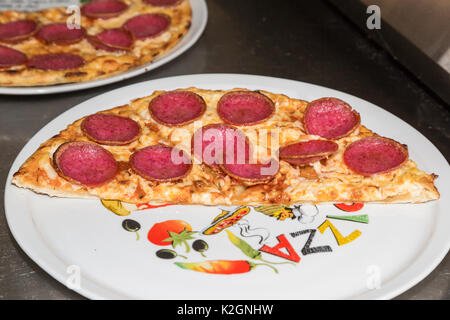 La metà di un caldo preparate di fresco pizza con salame, verdure e formaggio grattugiato da una stufa su una ceramica piastra bianca con una immagine dipinta in restaur Foto Stock