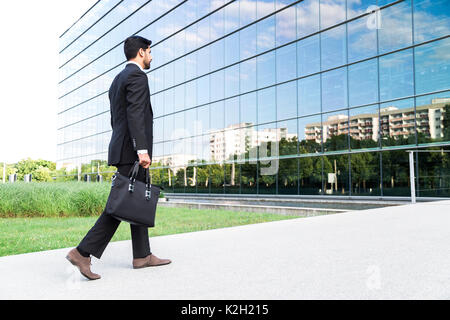 Anonimo imprenditore o lavoratore in abito nero con inserti in pelle borsa o custodia per il notebook in mano va a lavorare sulla strada che porta all'entrata di un vetro offi Foto Stock