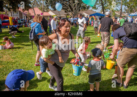 Multirazziale bambini che portano ceste sciame attraverso il prato di un parco in Costa Mesa, CA, ricerca di dispersi premi pasquali come una madre porta il suo bambino in una mano e i suoi premi nell'altro. Foto Stock