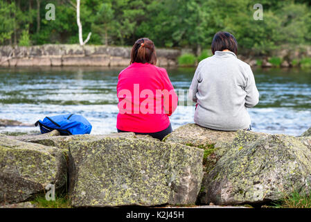 Setesdal, Norvegia - Agosto 16, 2017: documentario di viaggio di due donne seduti sulle pietre di fiume avente un talk, retrocede verso la telecamera. Fiume e f Foto Stock