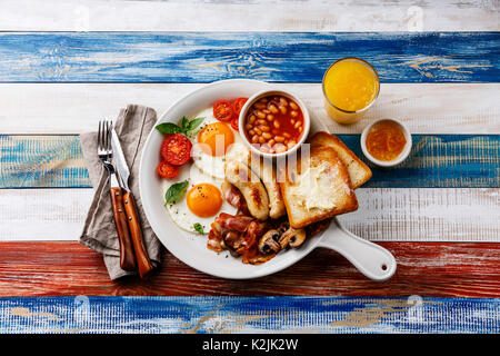 Prima colazione inglese in bianco padella con uova fritte, salsicce, pancetta, fagioli, toasts e fresco di colore arancione su sfondo di legno Foto Stock