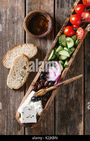 Ingredienti per il tradizionale insalata greca. Pomodori ciliegini, tagliati a fette cetrioli, cipolla rossa, olive nere, il formaggio feta in legno d'olivo ciotola con pane, o Foto Stock