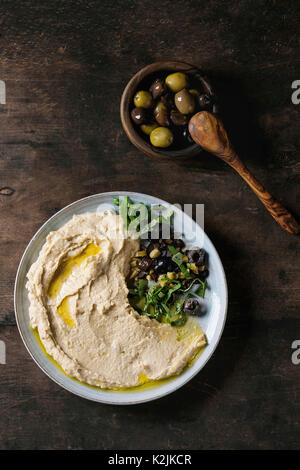 In casa tradizionale diffusione hummus con trinciatura di olive ed erbe sulla piastra blu, servito con una ciotola di olive varietà e il cucchiaio di legno su legno vecchio bac Foto Stock