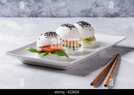 Mini riso sushi hamburger con salmone affumicato, insalata verde e salse, sesamo nero servito sul quadrato bianco piastra con bacchette di legno su cemento grigio Foto Stock