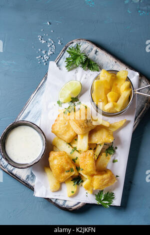 Tradizionale britannica fast food pesce e patatine. Servito con il formaggio bianco salsa, calce, prezzemolo, patatine fritte nel cestello sul libro bianco su blu co Foto Stock