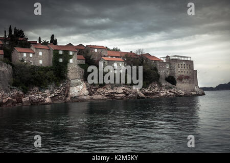 Vecchi edifici antichi su Sveti Stefan isola circondata da acqua di mare sulla costa durante il tramonto con cielo drammatico in Europa paese Montenegro Foto Stock