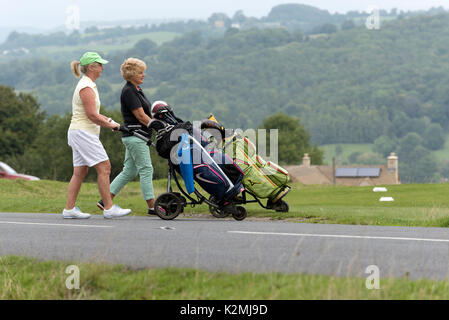 Signora golfisti nel sud Cotswolds regione del Regno Unito Inghilterra attraversando una strada di campagna con il loro campo da golf elettrico caddy. Auugust 2017 Foto Stock