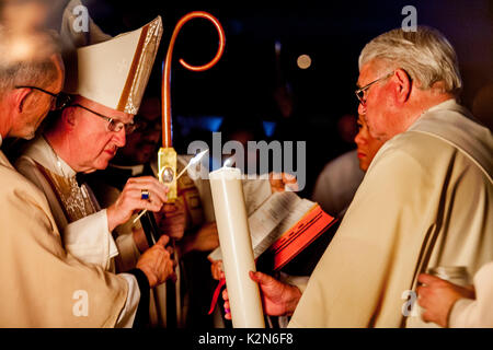 Dalla luce del fuoco pasquale, un vescovo cattolico utilizza un cono di luce di una candela processionale detenute da un diacono derubato prima di condurre una massa di battesimo presso un Orange, CA, Cattedrale. Foto Stock