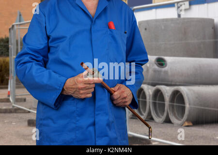 Plumber in tute blu da taglio di un tubo in rame mentre in piedi di fronte ad un mucchio di calcestruzzo tubazioni di drenaggio su un sito in costruzione Foto Stock