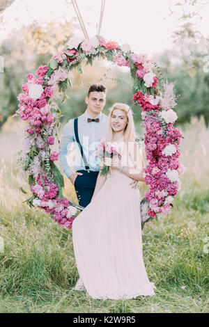 La foto verticale della damigella con il mazzo di fiori di colore rosa seduta sul matrimonio peonie arch e il miglior uomo in tuta blu in piedi dietro di lei. La composizione della foresta. Foto Stock
