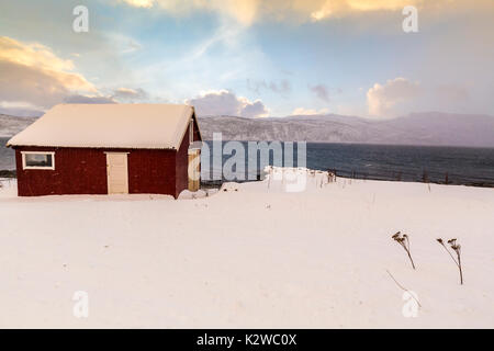 Tipici norvegesi calda e accogliente casa situata sulla riva del lago a un fiordo nel Troms County, Norvegia. Il sole è impostata bassa sopra l'orizzonte ed il cielo è Foto Stock