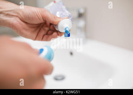 Le mani umane applicando una pasta dentifricia su un blu elettrico spazzolino. vi è un bianco una bacinella visibile sullo sfondo. dentifricio è graziosamente blu e fre Foto Stock