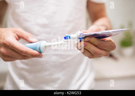 Close up di mani umane versando il dentifricio blu su uno spazzolino da denti elettrico. L uomo è sfocata, in una t-shirt bianco. Ci sono bagno visibile sul backgro Foto Stock