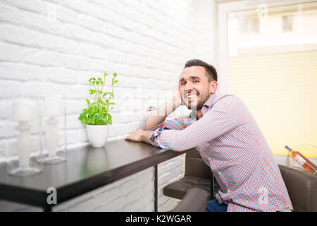 Giovane uomo seduto in cucina e sorridente Foto Stock