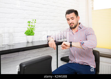 Giovane uomo che guarda il suo orologio con Viso confuso. Lui indossa camicia a scacchi e seduta in cucina Foto Stock