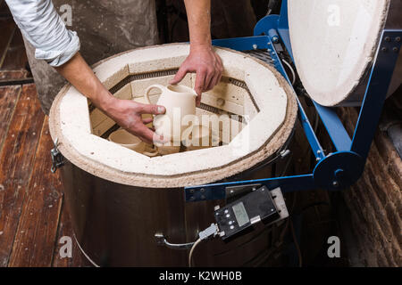 Ceramiche, workshop, arte ceramica concetto - maschio mani inserire delicatamente la caraffa nel forno, la vista in pianta del forno elettrico per ulteriore tostatura di argilla crudi pr Foto Stock