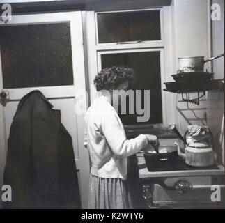 Anni sessanta, storiche, l'immagine di una donna per la cottura sul suo proprio su una stufa a gas in una piccola e semplice cucina sul retro di una casa a schiera, Inghilterra, Regno Unito. Foto Stock