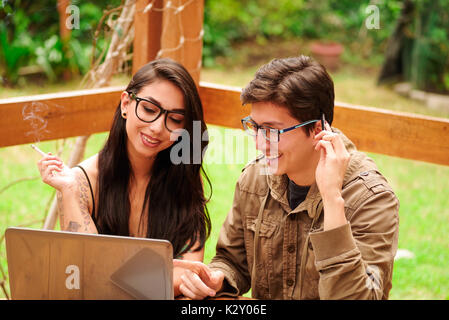 Bel giovane hipster sorridere mentre la sua ragazza è fumare una sigaretta, giovane che lavorano nel loro computer sul portico di casa in legno, estate rilassante concetto insieme in un cortile sullo sfondo Foto Stock