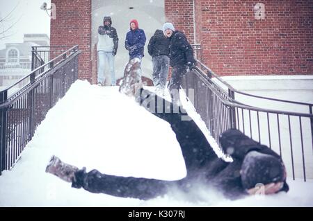 Uno studente scorre verso il basso una scale innevato con le gambe in aria e dei bracci che copre la sua testa, mentre gli altri studenti in marcia sulla neve il sorriso e allegria in cima alle scale, in un giorno di neve alla Johns Hopkins University, Baltimora, Maryland, 2016. Foto Stock