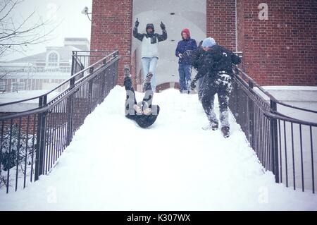 Uno studente scorre all'indietro con le gambe in aria e le braccia intorno alla sua testa, giù per una scalinata nevoso mentre gli amici in marcia sulla neve allietare dalla sommità delle scale, durante una giornata sulla neve alla Johns Hopkins University, Baltimora, Maryland, 2016. Foto Stock