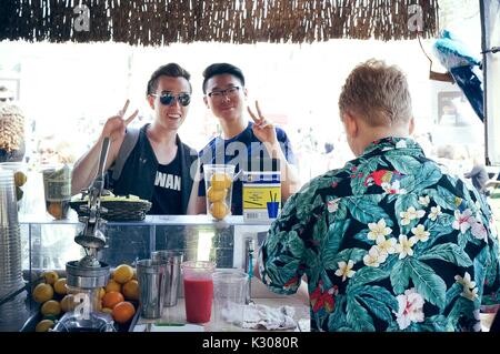 Due studenti pongono con segni di pace da dietro il bancone di un tiki-style lemonade stand, mentre un uomo in camicia hawaiana prepara le loro bevande, durante la Fiera di Primavera, uno studente-eseguire il carnevale di primavera presso la Johns Hopkins University, Baltimora, Maryland, aprile 2016. La cortesia Eric Chen. Foto Stock