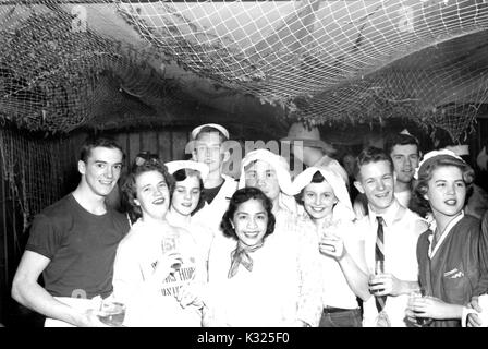 Durante un naufragio parte ospitata da Delta Upsilon fraternità alla Johns Hopkins University, sorridente maschio e femmina fêtards pongono insieme azienda bevande e indossando abiti nautico, sotto un soffitto coperto in un mare net, Baltimore, Maryland, 1953. Foto Stock