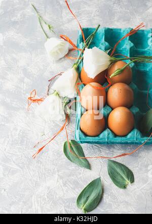 Uova crude in una scatola di cartone per le uova con fiori di colore bianco su un marmo bianco da tavola, pronto per la verniciatura di Pasqua, lo spazio al di sotto del testo, il fuoco selettivo Foto Stock