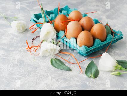 Uova crude in una scatola di cartone per le uova con fiori di colore bianco su un marmo bianco da tavola, pronto per la verniciatura di Pasqua, lo spazio al di sotto del testo, il fuoco selettivo Foto Stock