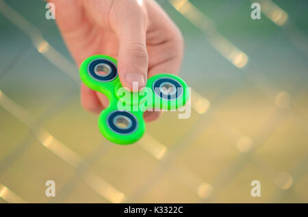 Giovani man mano che regge il gadget antistress agitano: spinner. Adolescente giocando con il filatore verde all'aperto sulla griglia rabitz bokeh di fondo. DOF poco profondo. Foto Stock