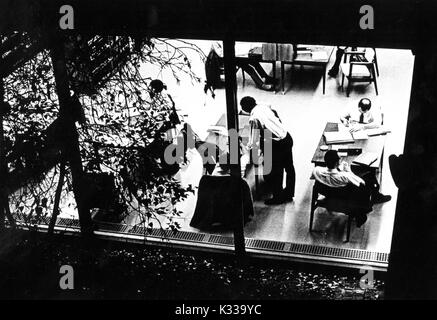 Attraverso le finestre di vetro che conducono fuori per gli alberi e gli arbusti sul cortile di Milton S Eisenhower biblioteca presso la Johns Hopkins University, studenti indossando abiti lavoro a tabelle all'interno della libreria sul livello M, seduto di fronte a libri di testo e di pile di carta, con un paio di studenti a conversare in corrispondenza di un'altra tabella, Baltimore, Maryland, 1965. Foto Stock