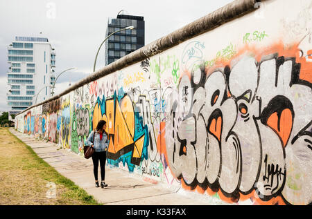 Giovane donna camminare passato graffiti sul muro di Berlino, la East side gallery, muhlenstrasse, quartiere Friedrichshain di Berlino, Germania, Europa Foto Stock