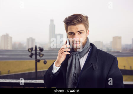 La foto di un giovane imprenditore in piedi da qualche parte al di fuori contro di background urbano. Egli sta parlando al cellulare. Foto Stock
