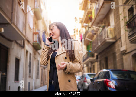 Una foto di una giovane donna in cappotto beige in piedi in mezzo alla strada e parlare al cellulare. Lei è sorridente. Foto Stock