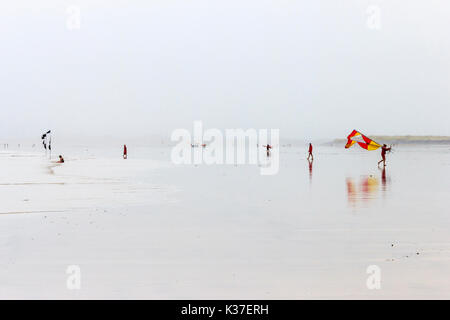 Bagnino RNLI portando il giallo e il rosso flag di avviso e surfisti su un tetro spiaggia sabbiosa a bassa marea in una piovosa e ventoso giorno in estate Foto Stock