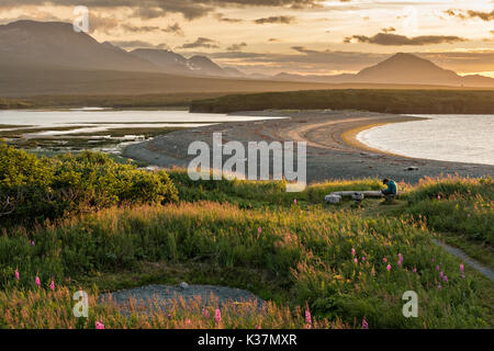 Un camper orologi il tramonto sopra le montagne Chigmit e McNeil Cove al McNeil River State Game Santuario sulla Penisola di Kenai, Alaska. Il sito remoto è accessibile solo con un permesso speciale ed è il più grande del mondo di popolazione stagionale di orsi bruni nel loro ambiente naturale. Foto Stock