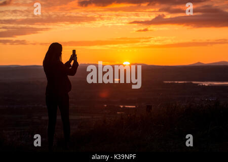 Una donna prende una foto del tramonto con il suo smart phone dal picco Coaley nel Gloucestershire guardando verso il fiume Severn e la Foresta di Dean. Foto Stock