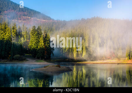 Lago di montagna con piccola isola riflette un ambiente di misty foreste di abete rosso su pendii. lo straordinario paesaggio con una fitta nebbia tra gli alberi a f Foto Stock