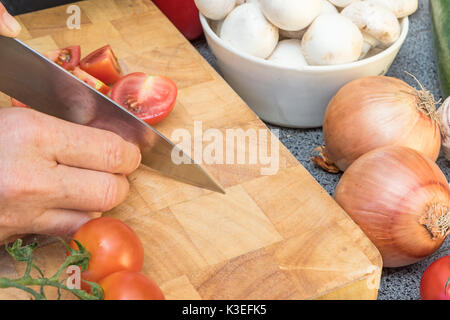 Vista laterale della donna per affettare un pomodoro su una tavola di legno. Vari tipi di verdure e funghi sono giacenti sul tavolo. Foto Stock