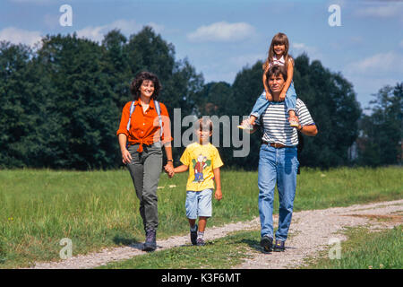 Gita di famiglia a piedi, madre detiene il figlio in mano, padre figlia porta sulle spalle Foto Stock