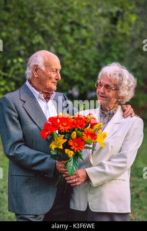 Senior Citizen's giovane con bouquet, l'uomo ha messo il suo braccio intorno alla donna e li guarda Foto Stock