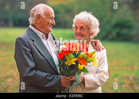 Senior Citizen's giovane con bouquet, l'uomo ha messo il suo braccio intorno a la donna e la guarda Foto Stock