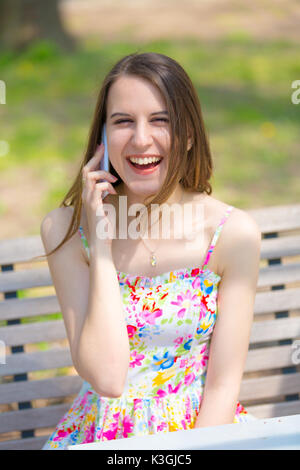 Ritratto di giovane bella donna con capelli lunghi in estate park, ragazza fiore di indossare abiti corti è utilizzando uno smartphone mentre è seduto su una panchina Foto Stock