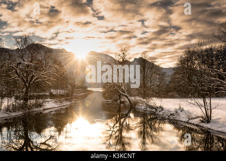 Il sole di sera sembra circa la silhouette delle prealpi bavaresi e viene riflessa nell'acqua del Kochelsee. Coperte di neve scenario della vecchia capanna in legno e gli alberi in primo piano. Foto Stock