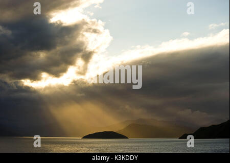 Sunray rompere attraverso una nube spessa copertura sopra il mare e illuminata di una piccola isola e in background di fiordi nel lato nord dell'isola del sud della Nuova Zelanda. Foto Stock