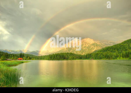 Doppio arcobaleno su northern Karwendelgebirge (montagne) su Mittenwald. La superficie dell'acqua dell'Lautersee (lago) è facilmente arricciato dall inizio pioggia. A sinistra nella foto è il tetto rosso di una piccola piscina esterna con chiosco sulla destra il riparo acqua piccola casa. Foto Stock